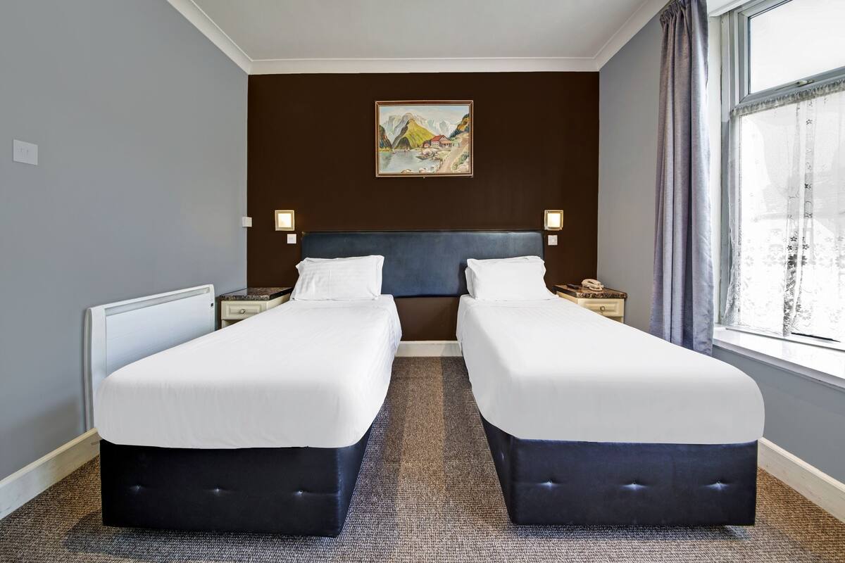 A hotel room with two beds, a large window with curtains, a painting on the wall, and a carpeted floor.