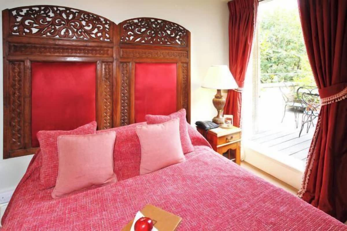 Ein Bett mit roter Kopfteil und Kissen, ein Nachttisch mit Lampe und Blick ins Freie.