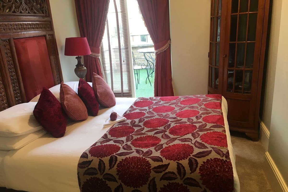 Ein Hotelzimmer mit Bett, rot-weißer Bettwäsche, einem Holz-Kopfteil, einer roten Lampe und Blick auf einen Außenbereich mit Tisch und Stühlen.