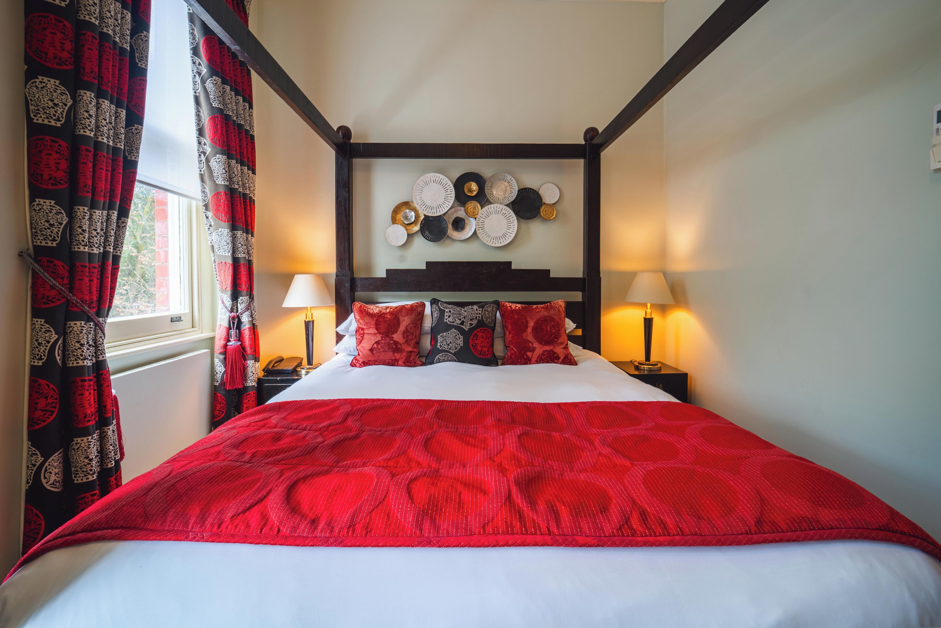 A four-poster bed with a red bedspread, two bedside lamps, and a wall-mounted plate collection.