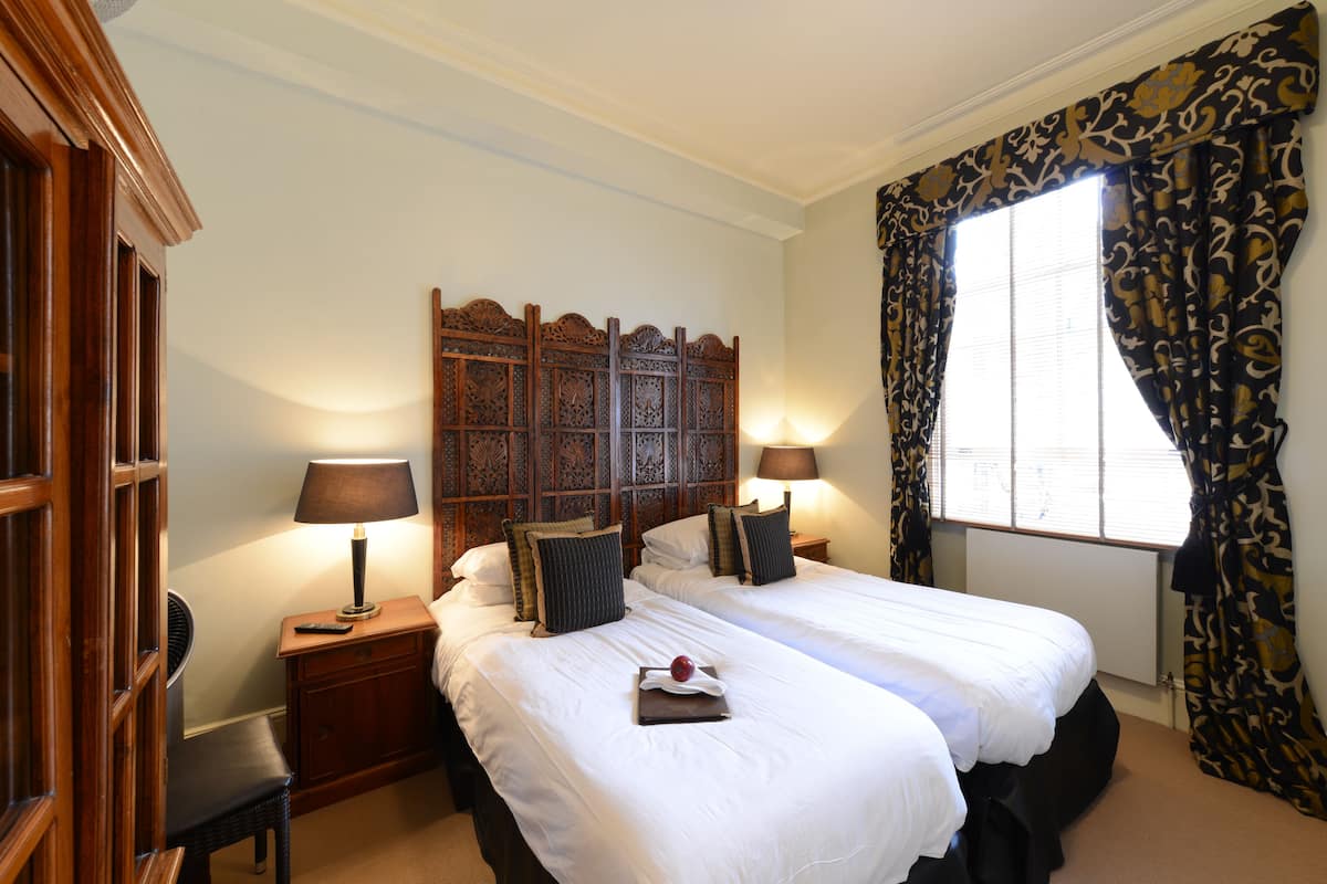 Ein Hotelzimmer mit zwei Betten, einem Holz-Kopfteil, Nachttischlampen, einem Fenster mit Vorhängen und einem kleinen Tisch mit Lampe.