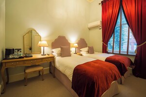 A hotel room with two beds, a desk with a chair, a mirror, and a window with red curtains.
