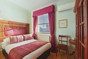 Ein ordentlich bezogenes Bett mit Kopfteil, ein Fenster mit Vorhängen, ein Stuhl und ein kleiner Tisch mit einer Schale.