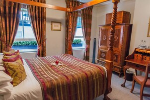 Ein Bett mit Baldachin, eine karierte Bettdecke, ein Nachttisch mit Lampe und Blick ins Freie durch ein Fenster.