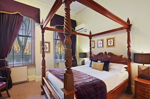 A four-poster bed with a wooden frame, a large window with blinds, a chair, and a nightstand with a lamp.