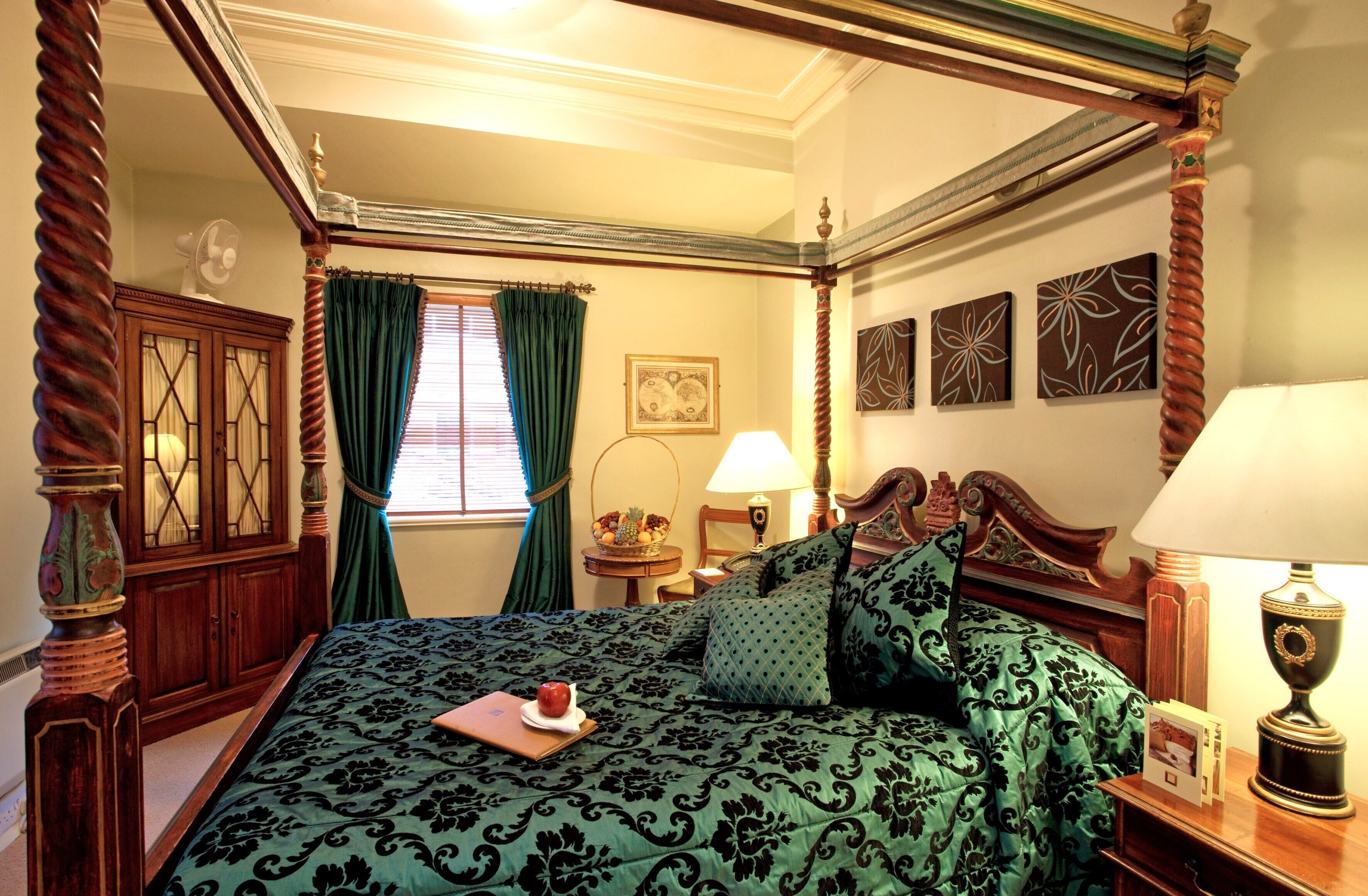 A four-poster bed with a green damask bedspread, a wooden nightstand with a lamp, a side table with a lamp, and a window with green curtains.