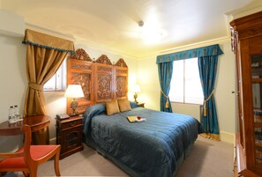 A bedroom with a large bed, a wooden nightstand with a lamp, a desk with a chair, and a wardrobe.