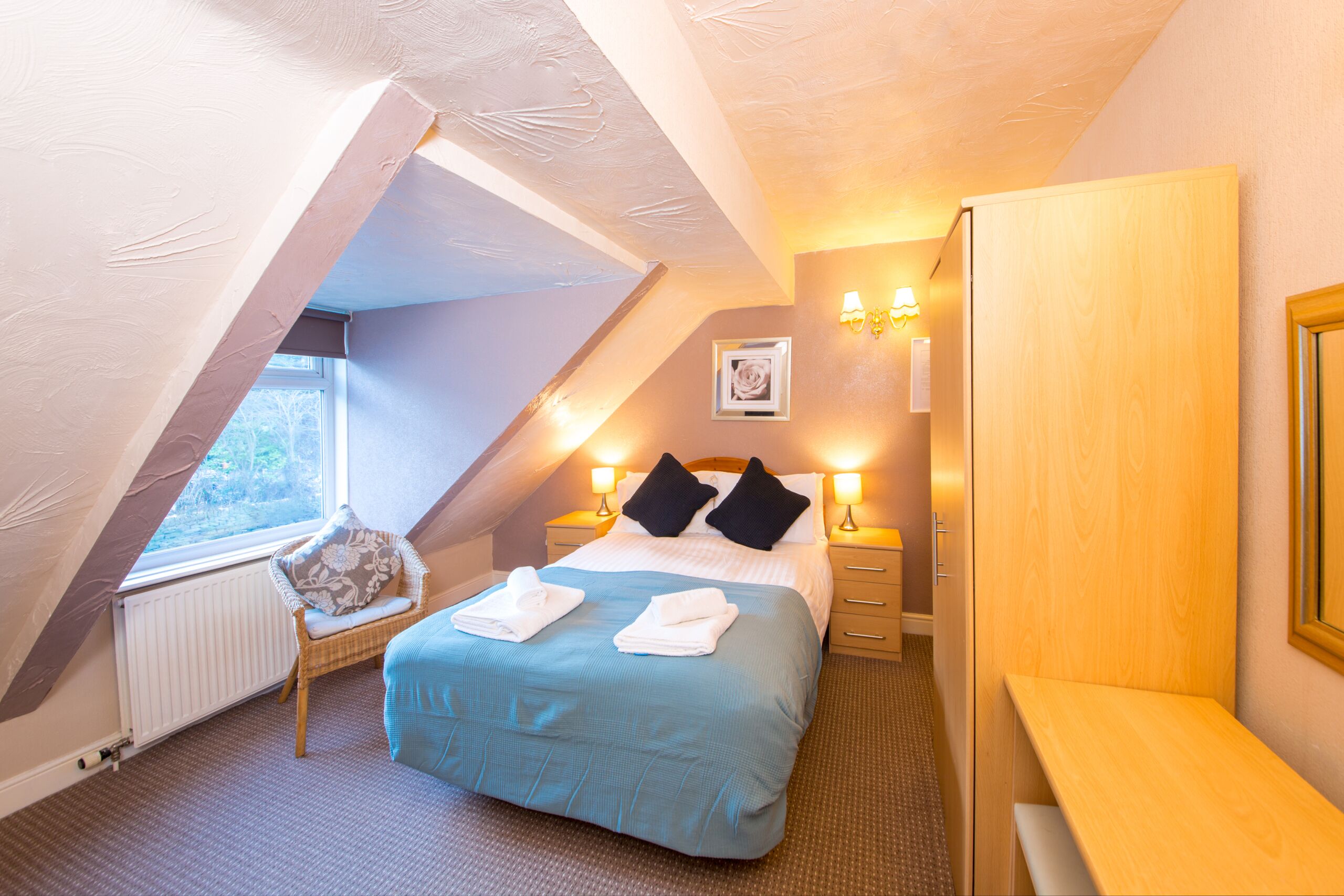 A bedroom with a sloped ceiling, a bed with a blue bedspread, two black pillows, a wooden nightstand with a lamp, and a window with a view of trees.