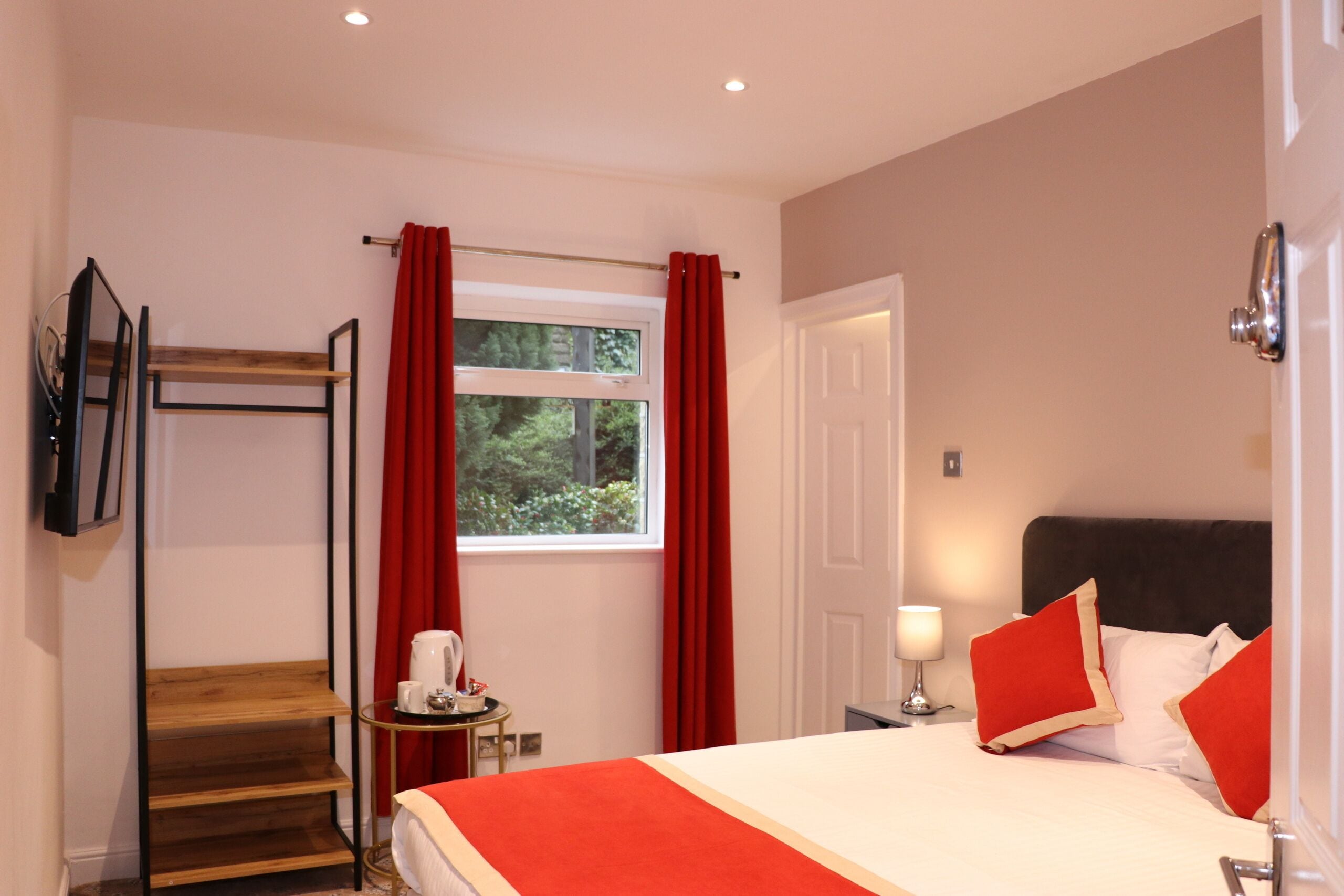 A bedroom with a bed, a wooden shelf, a small table with a lamp, and a window with red curtains.