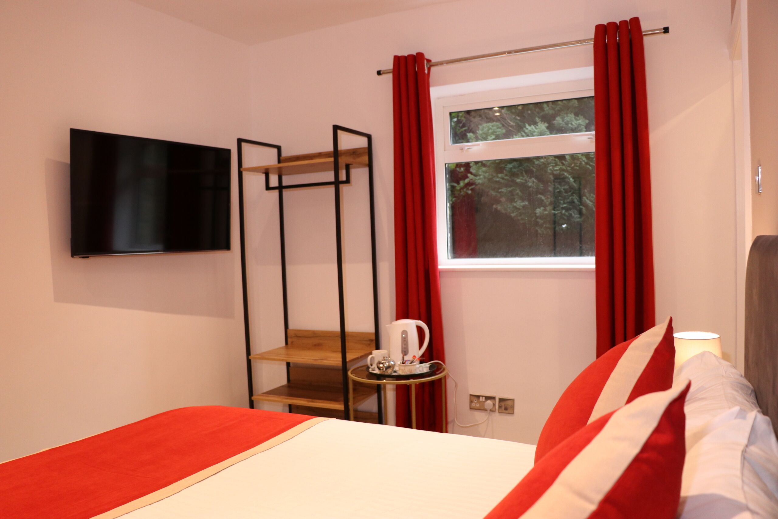 A hotel room with a bed, a TV mounted on the wall, a window with red curtains, and a small table with a kettle and cup.