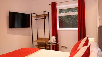 A hotel room with a bed, a TV mounted on the wall, a window with red curtains, and a small table with a kettle and cup.