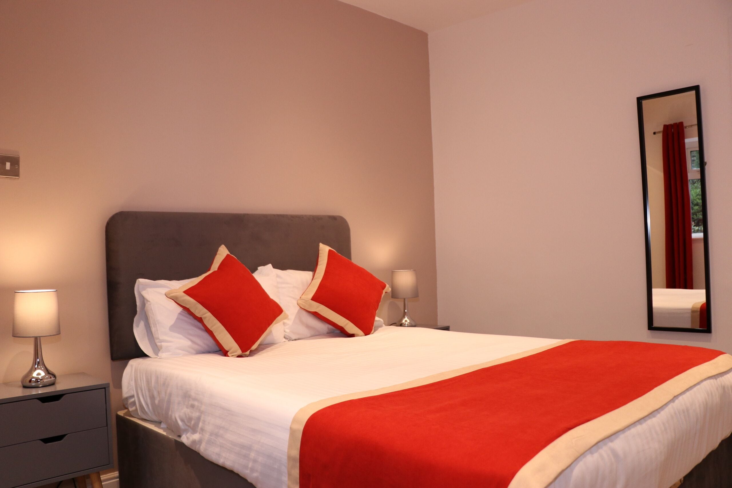 Un lit bien fait, avec du linge de lit blanc et des touches de rouge, une table de chevet avec une lampe et un miroir au mur.