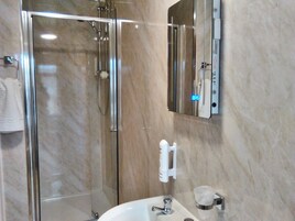 Un baño moderno con ducha envuelta en vidrio, un lavamanos blanco con grifo cromado y un espejo con reloj digital.