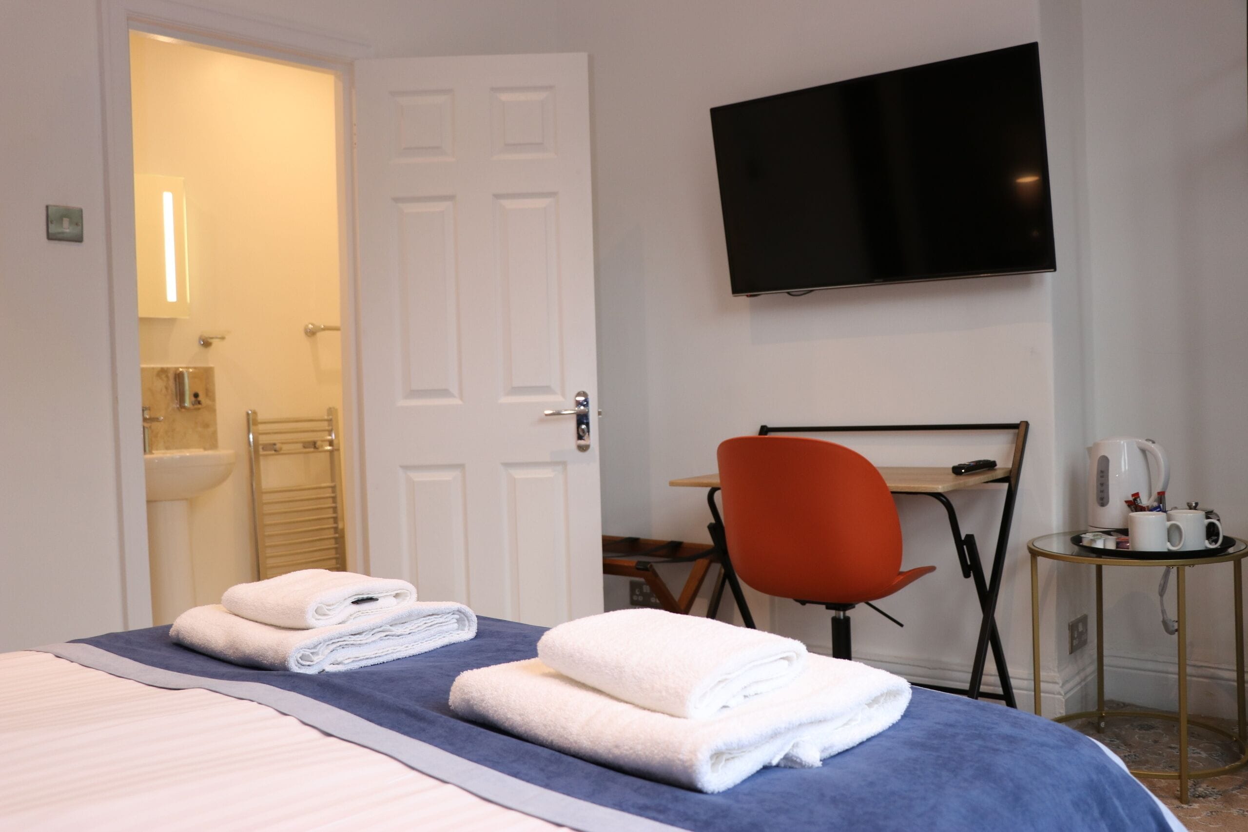 Habitación de hotel con una cama, un televisor montado en la pared, un escritorio con una silla y una mesita con hervidor y tazas.