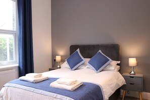 A neatly made bed with blue and white bedding, two pillows, and two folded towels. There are bedside lamps on either side of the bed. - Dalesgate Hotel (Keighley)