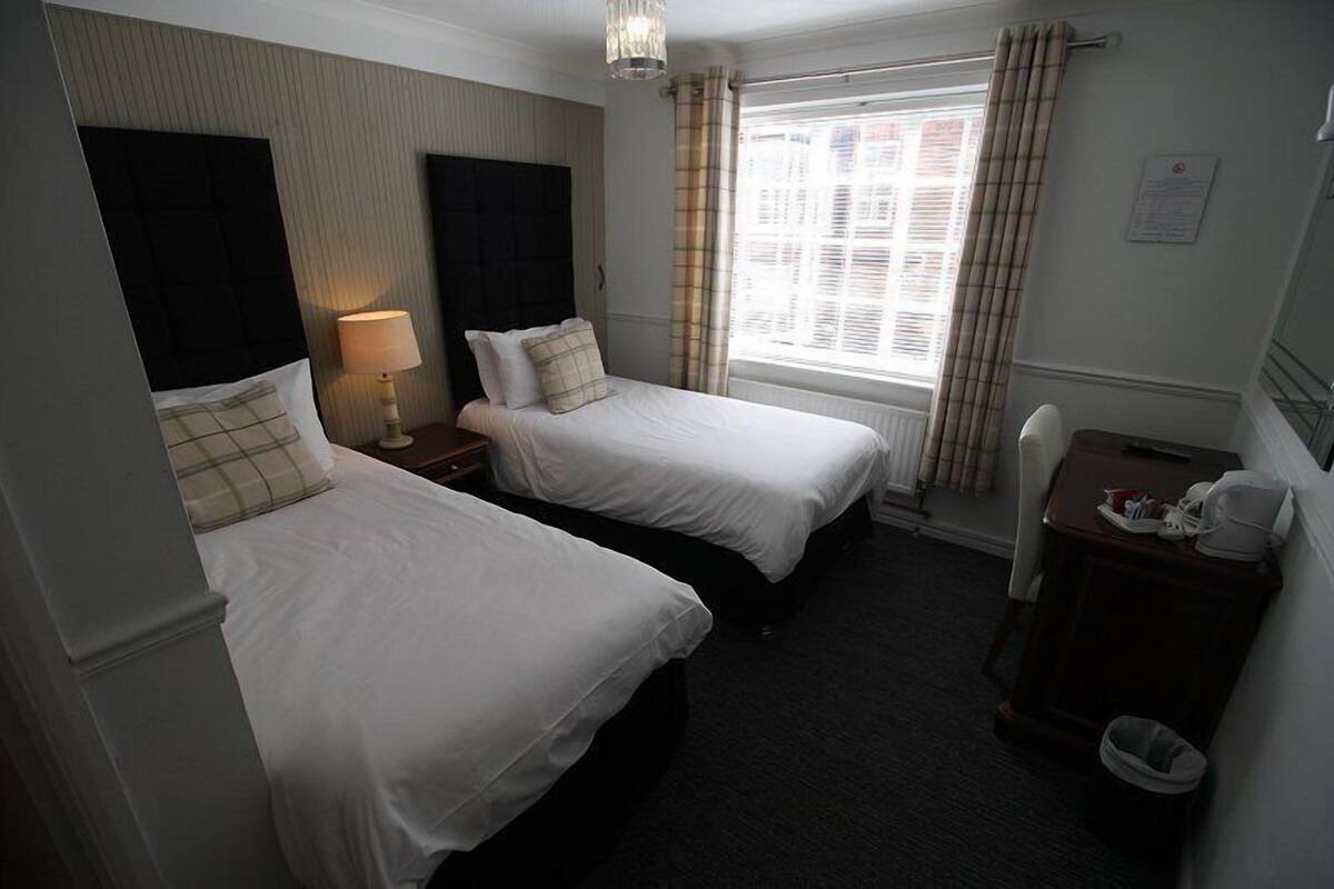 A hotel room with two single beds, a small desk with a chair, a lamp, and a window with curtains.