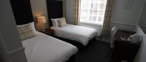 A hotel room with two single beds, a small desk with a chair, a lamp, and a window with curtains.