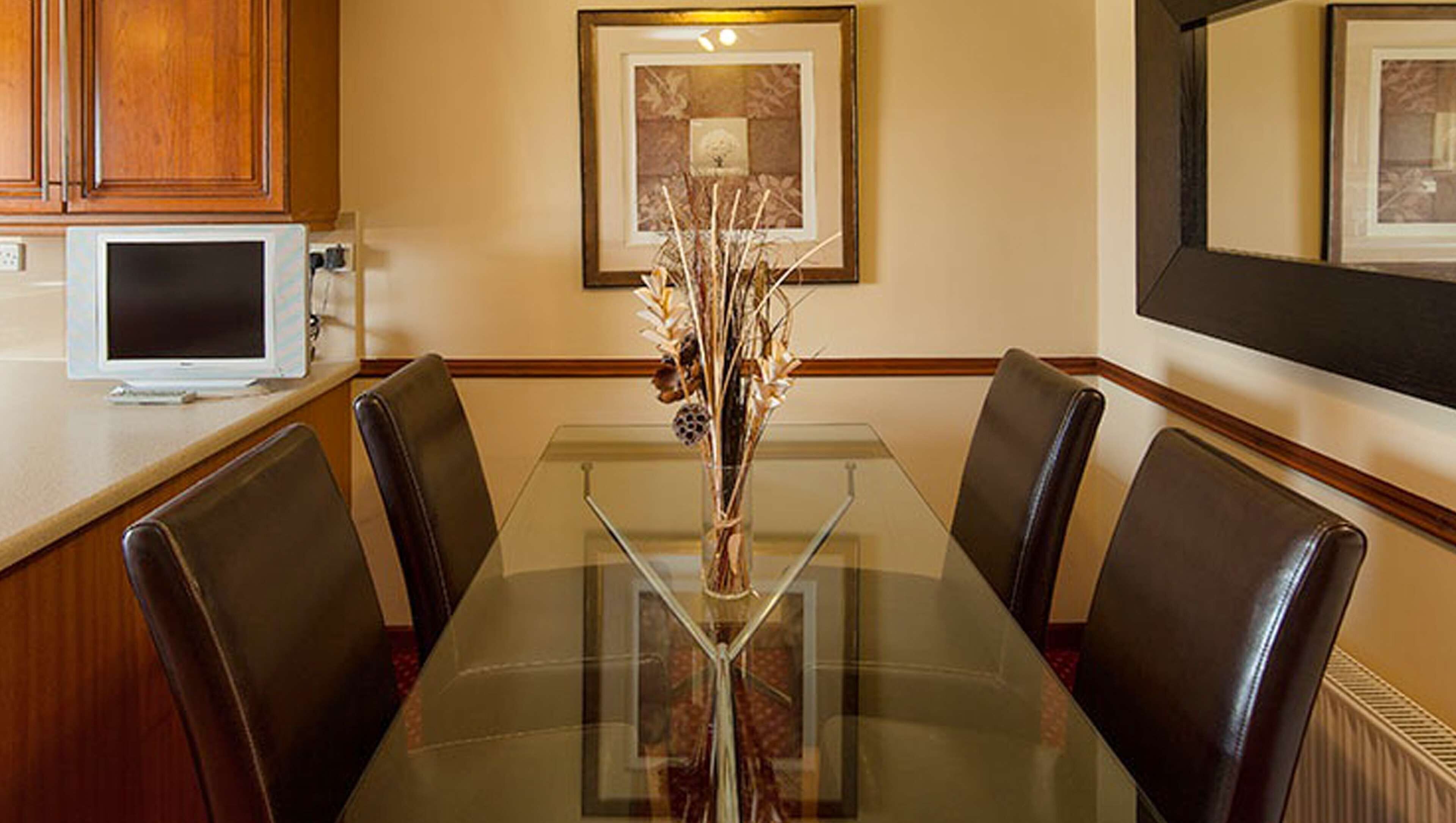 A dining area with a glass table surrounded by leather chairs, a framed artwork on the wall, and a white appliance on the counter.
