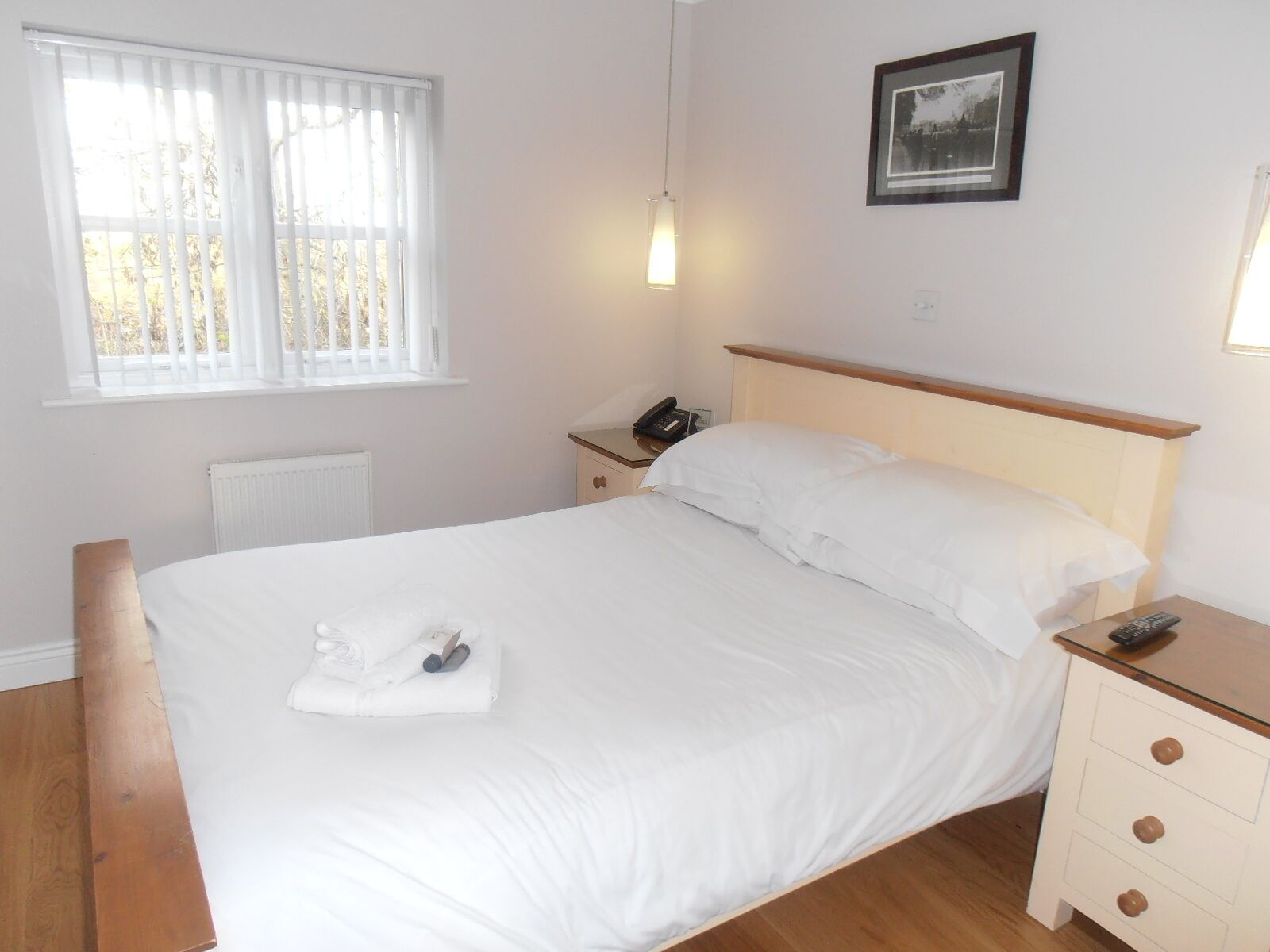 A neatly made bed with white linens, a bedside table with a lamp, a framed picture on the wall, and a window with blinds.