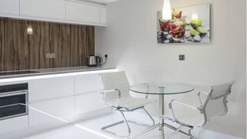 Uma cozinha moderna com armários brancos, uma chaleira de aço inoxidável, uma mesa de vidro com cadeiras de metal e uma obra de arte na parede com tema de frutas.