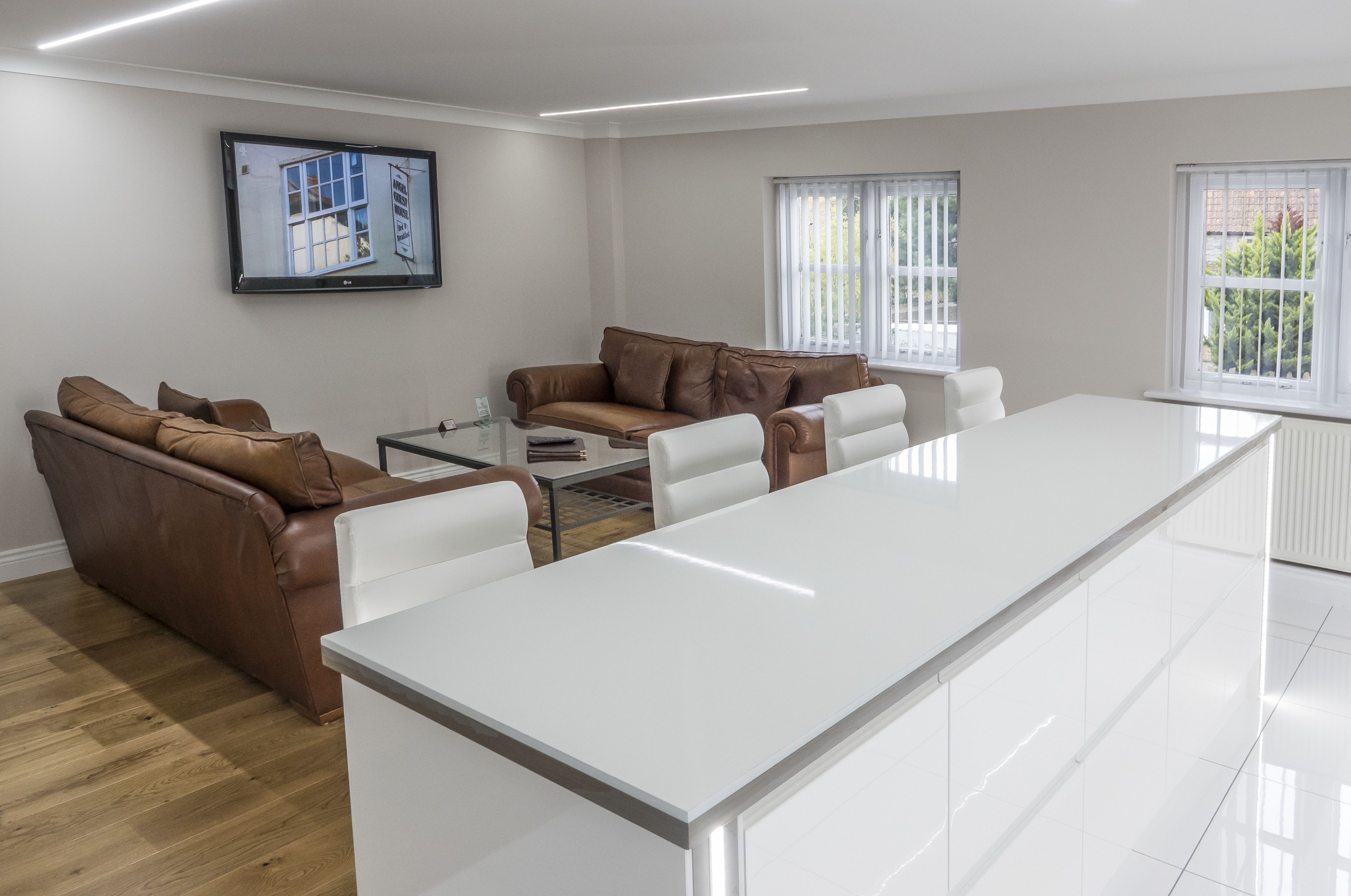 A modern living room with a long white table, brown leather sofas, and a flat-screen TV on the wall.