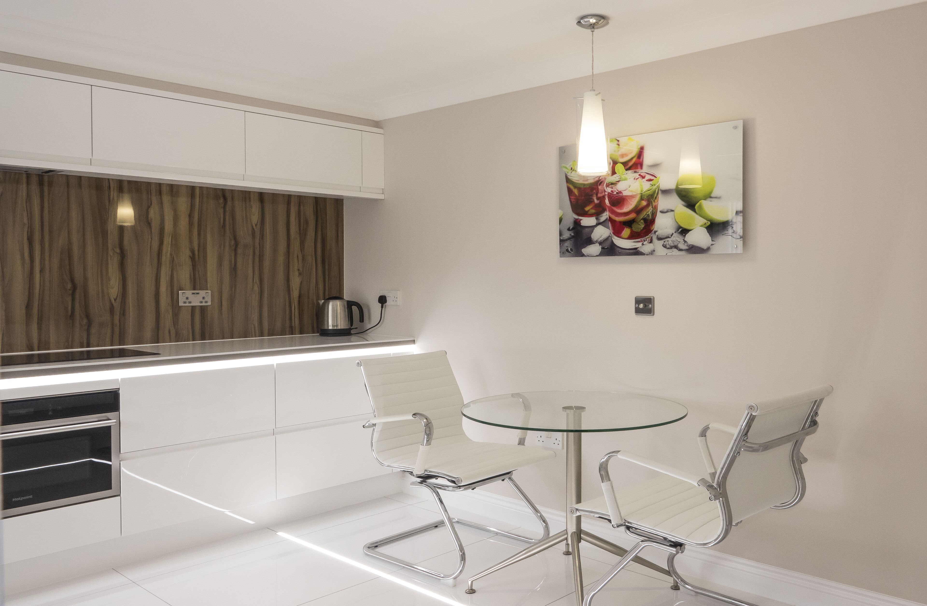 A modern kitchen with white cabinets, a wooden accent wall, a glass dining table with two chairs, and a fruit-themed artwork on the wall.