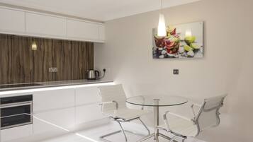 A modern kitchen with white cabinets, a wooden accent wall, a glass dining table with two chairs, and a fruit-themed artwork on the wall.