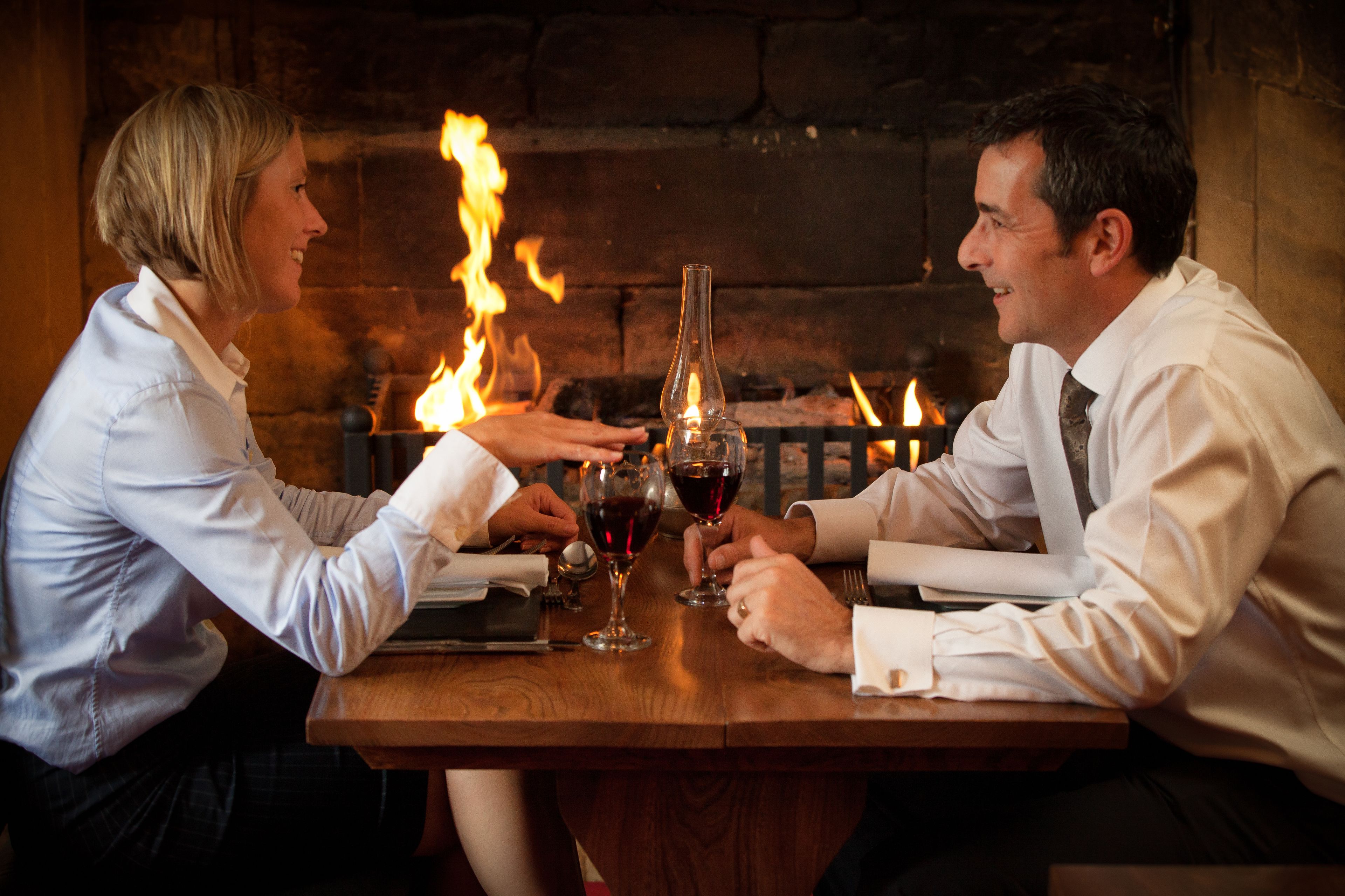 A couple seated at a wooden table with a bottle and glasses, a fireplace with flames in the background.