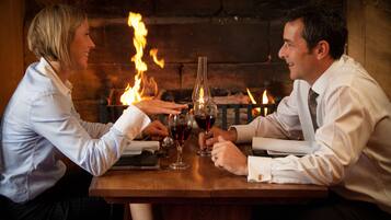 A couple seated at a wooden table with a bottle and glasses, a fireplace with flames in the background.