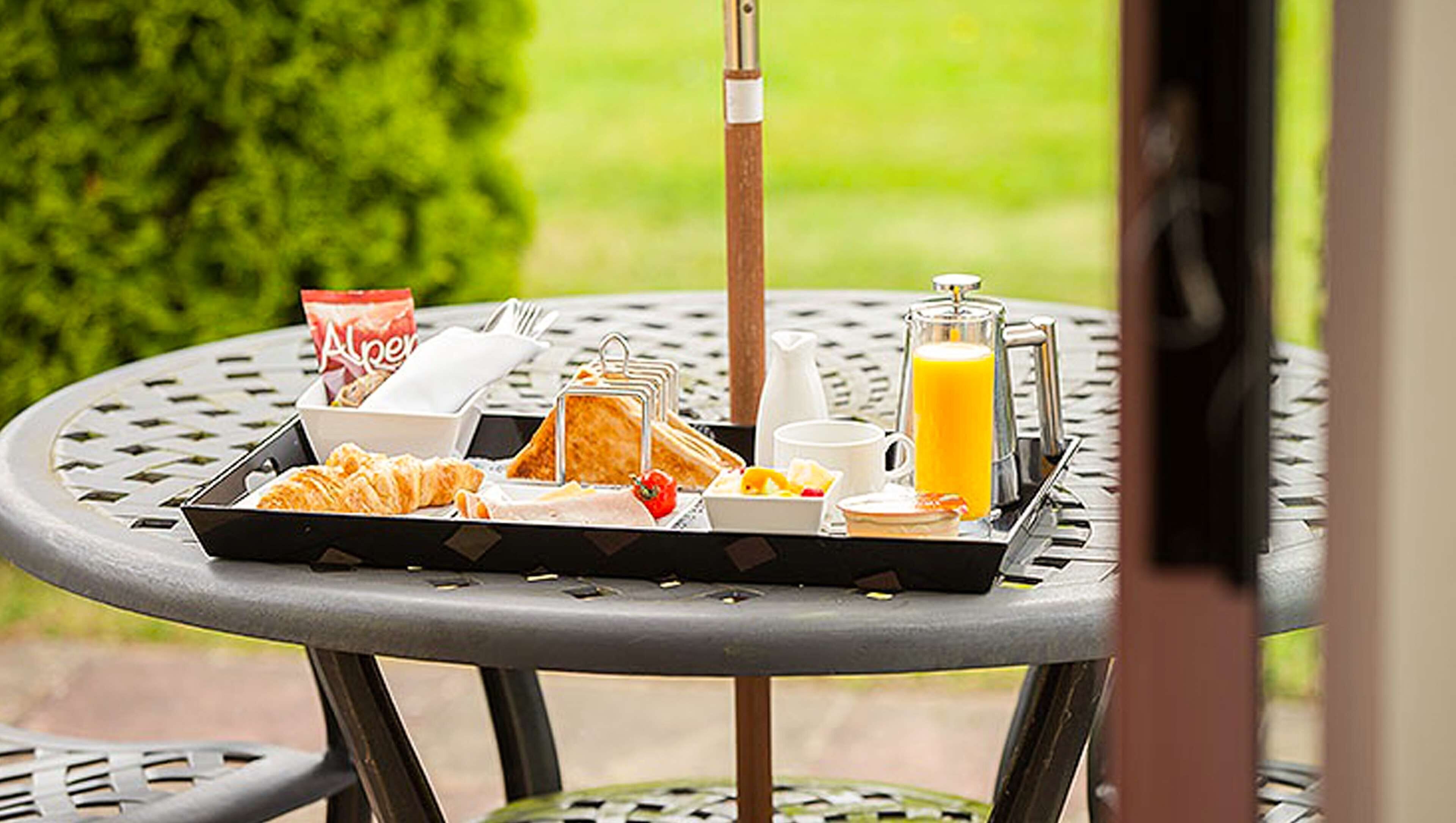En bricka med frukost på ett utomhusbord, innehållande croissanter, en flaska juice, ett glas apelsinjuice, en kopp och ett paket socker.