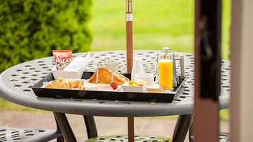 En bricka med frukost på ett utomhusbord, innehållande croissanter, en flaska juice, ett glas apelsinjuice, en kopp och ett paket socker.