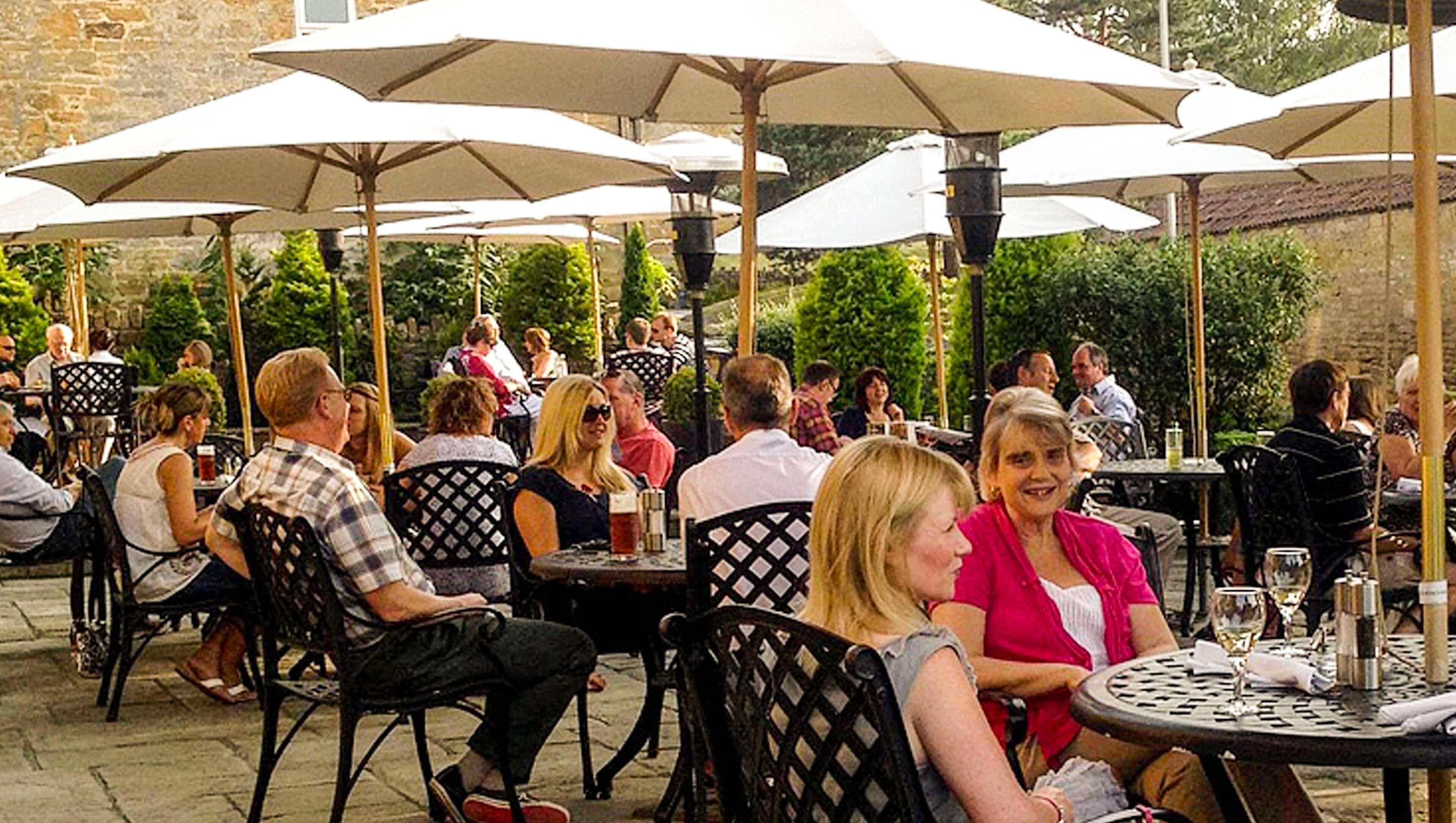 An outdoor seating area with tables and chairs under umbrellas, surrounded by greenery and stone walls.