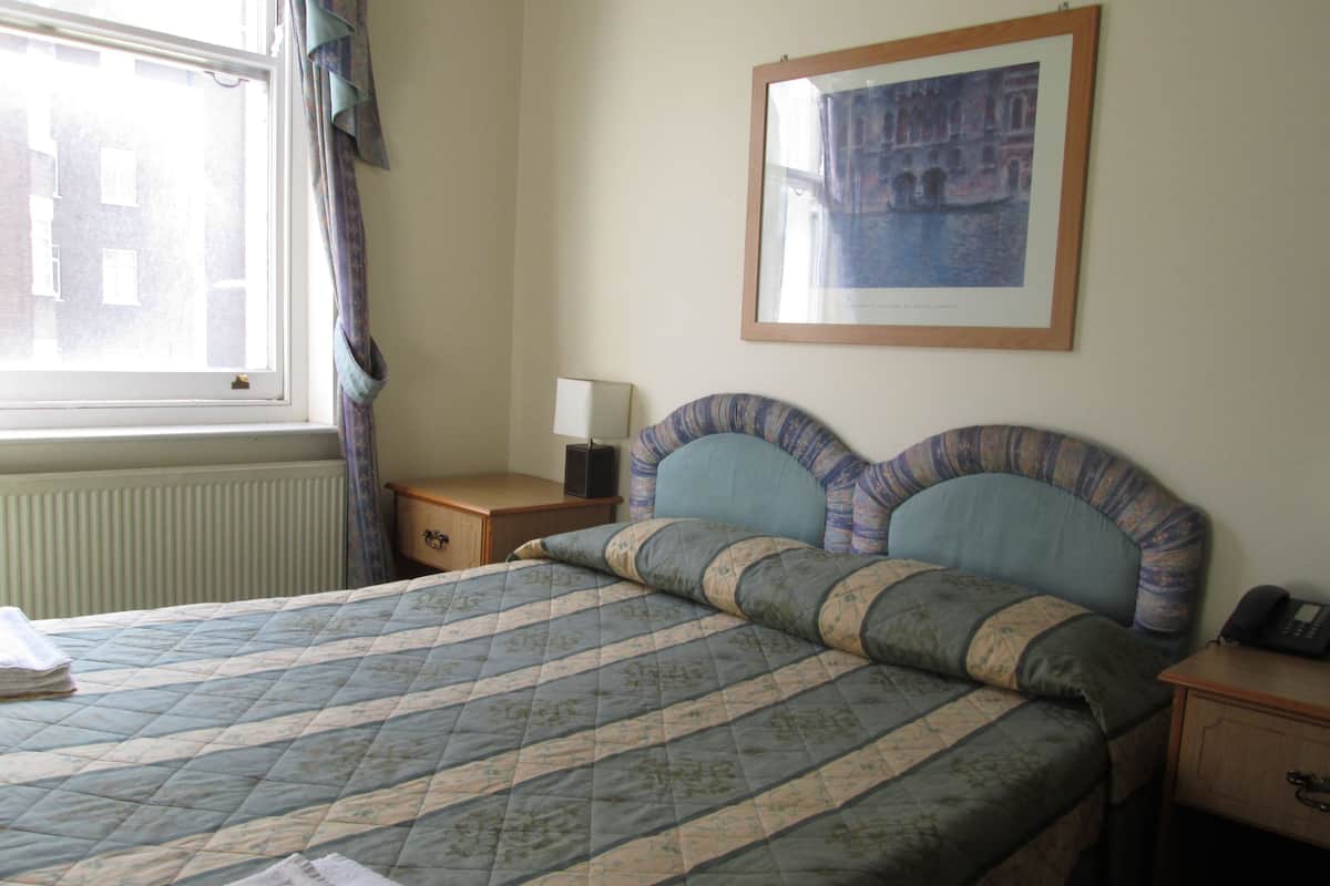 A hotel room with a bed, a bedside table with a lamp, a framed picture on the wall, and a window with curtains.
