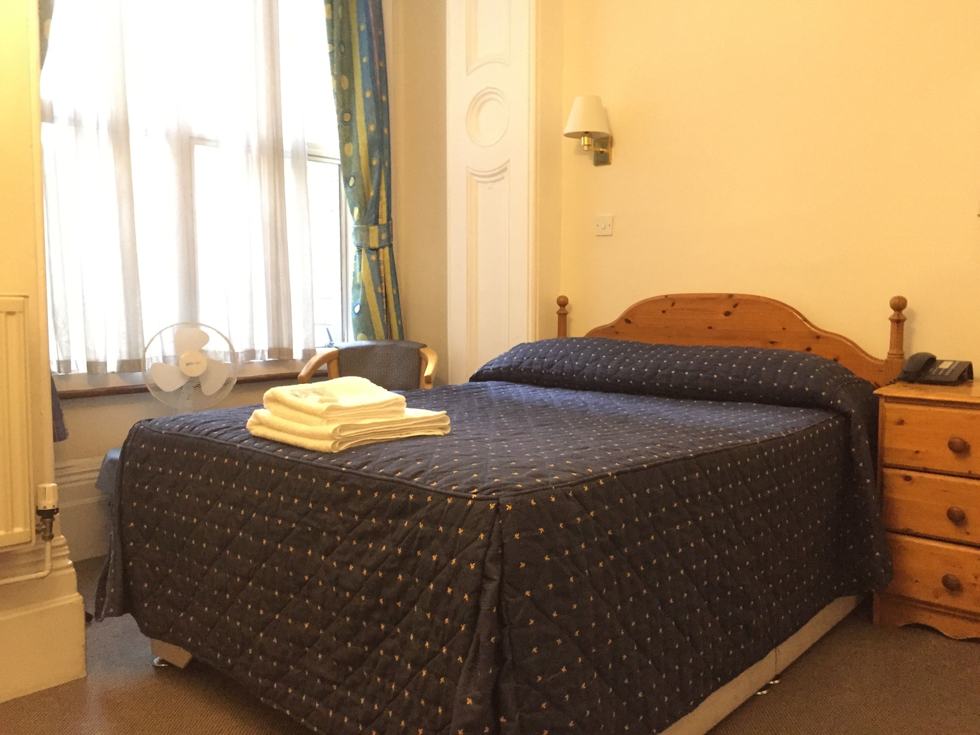 A neatly made bed with a dark quilt, a folded towel on the footboard, a bedside lamp, and a nightstand with a phone.