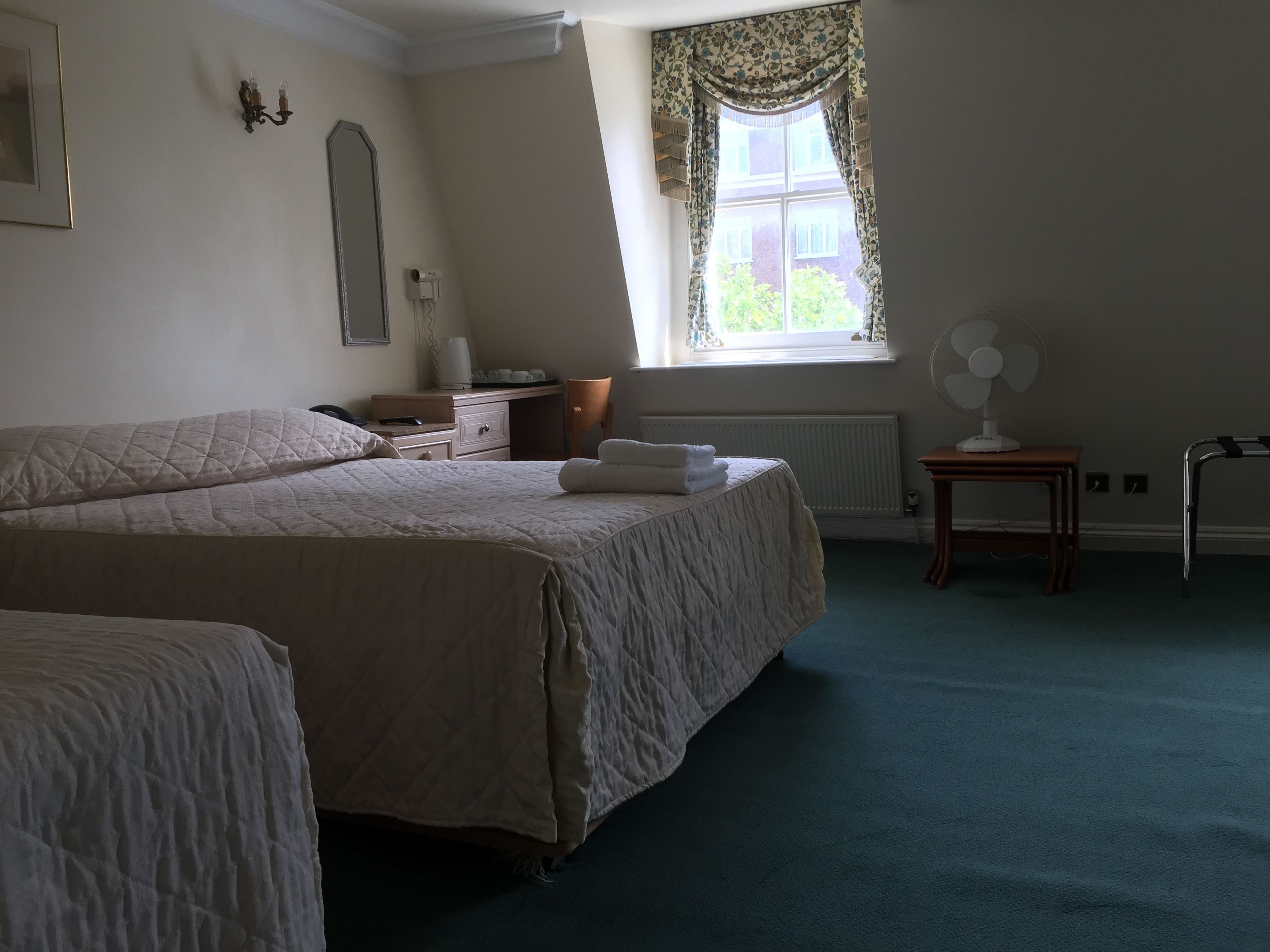 A bedroom with two beds, a bedside table, a lamp, a radiator, a window with curtains, and a standing fan.