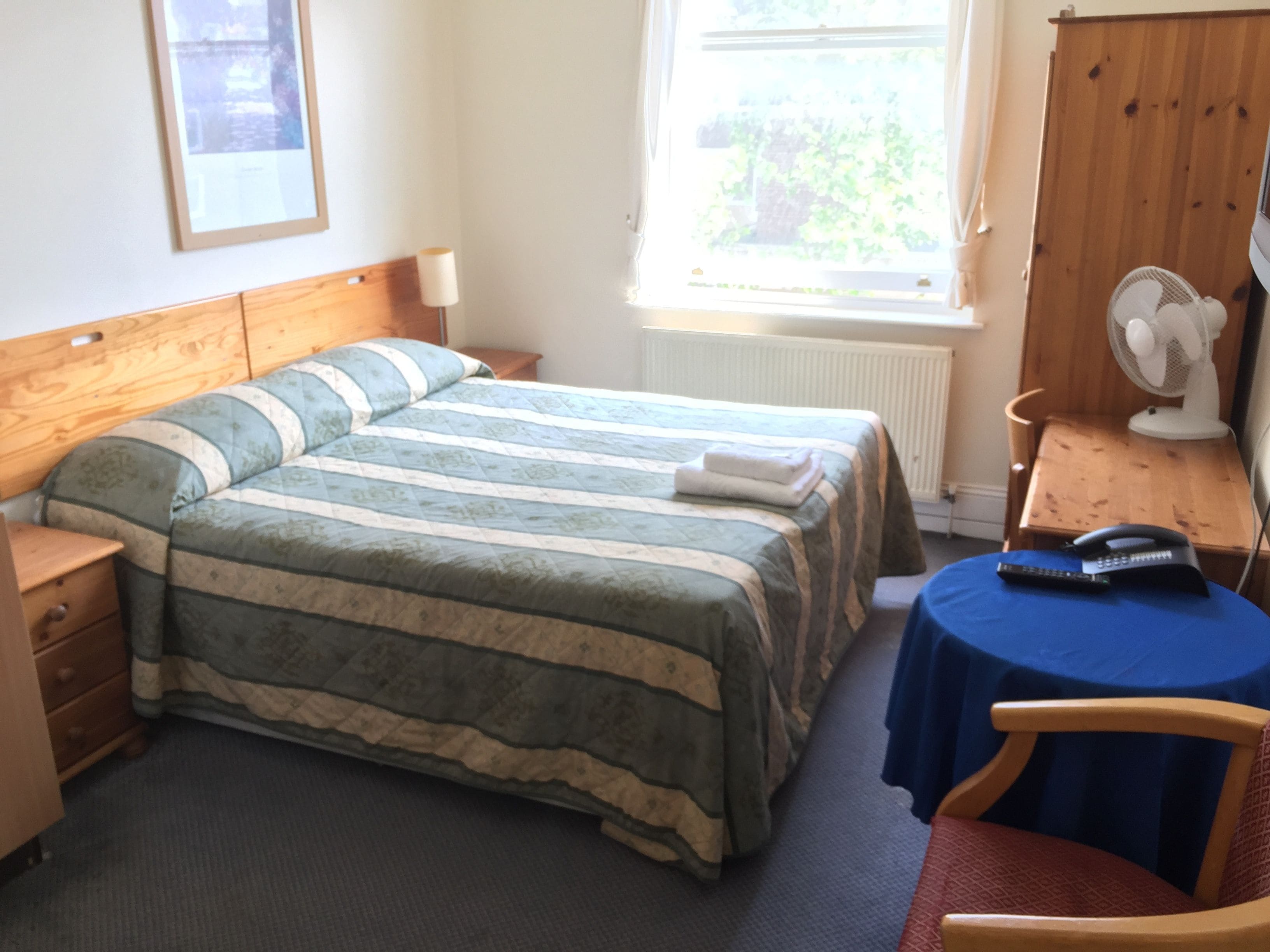 A bedroom with a bed, a bedside table, a chair, a desk, a fan, and a window.