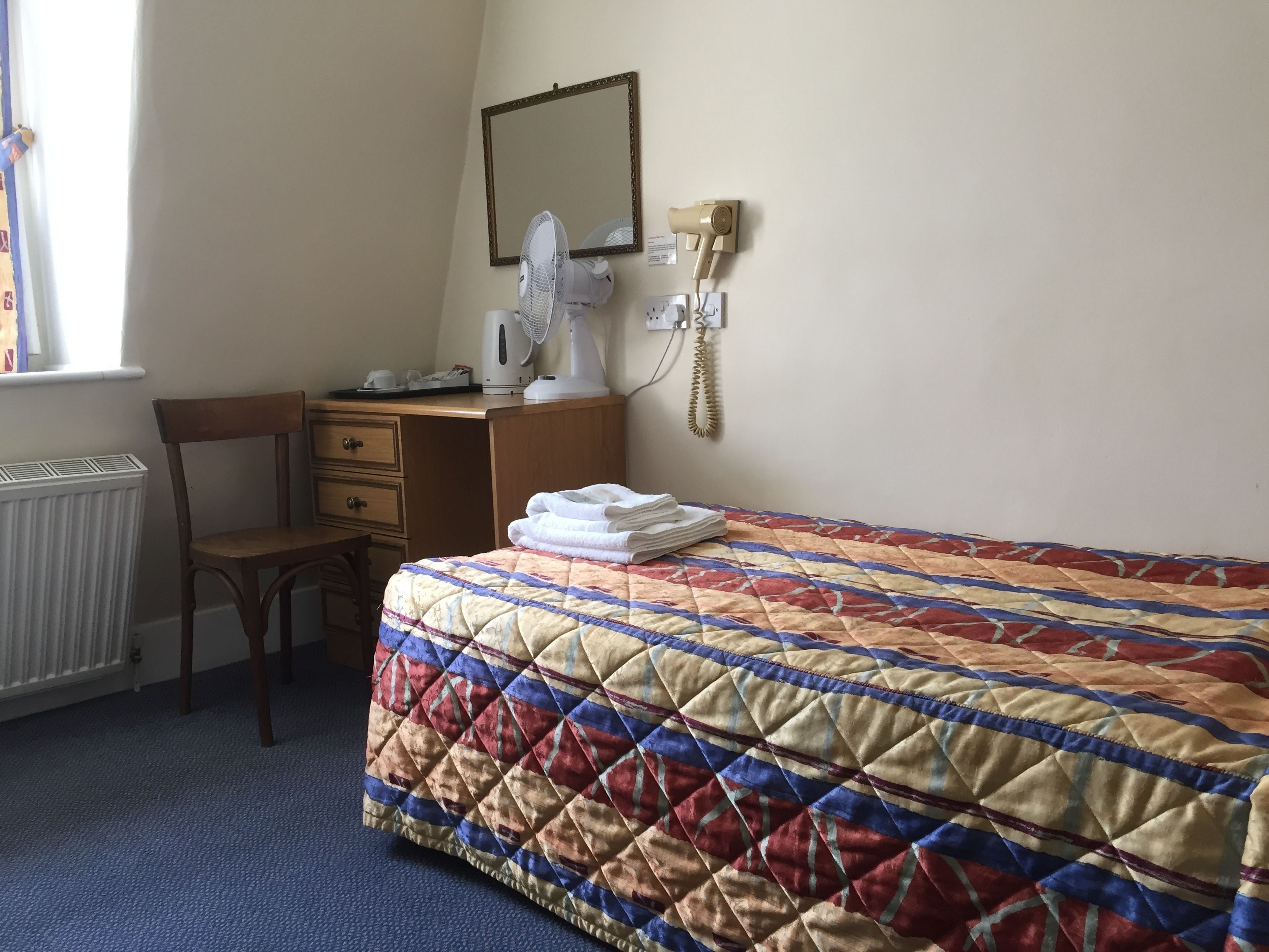 A hotel room with a bed, a chair, a desk, a mirror, a radiator, and a telephone.