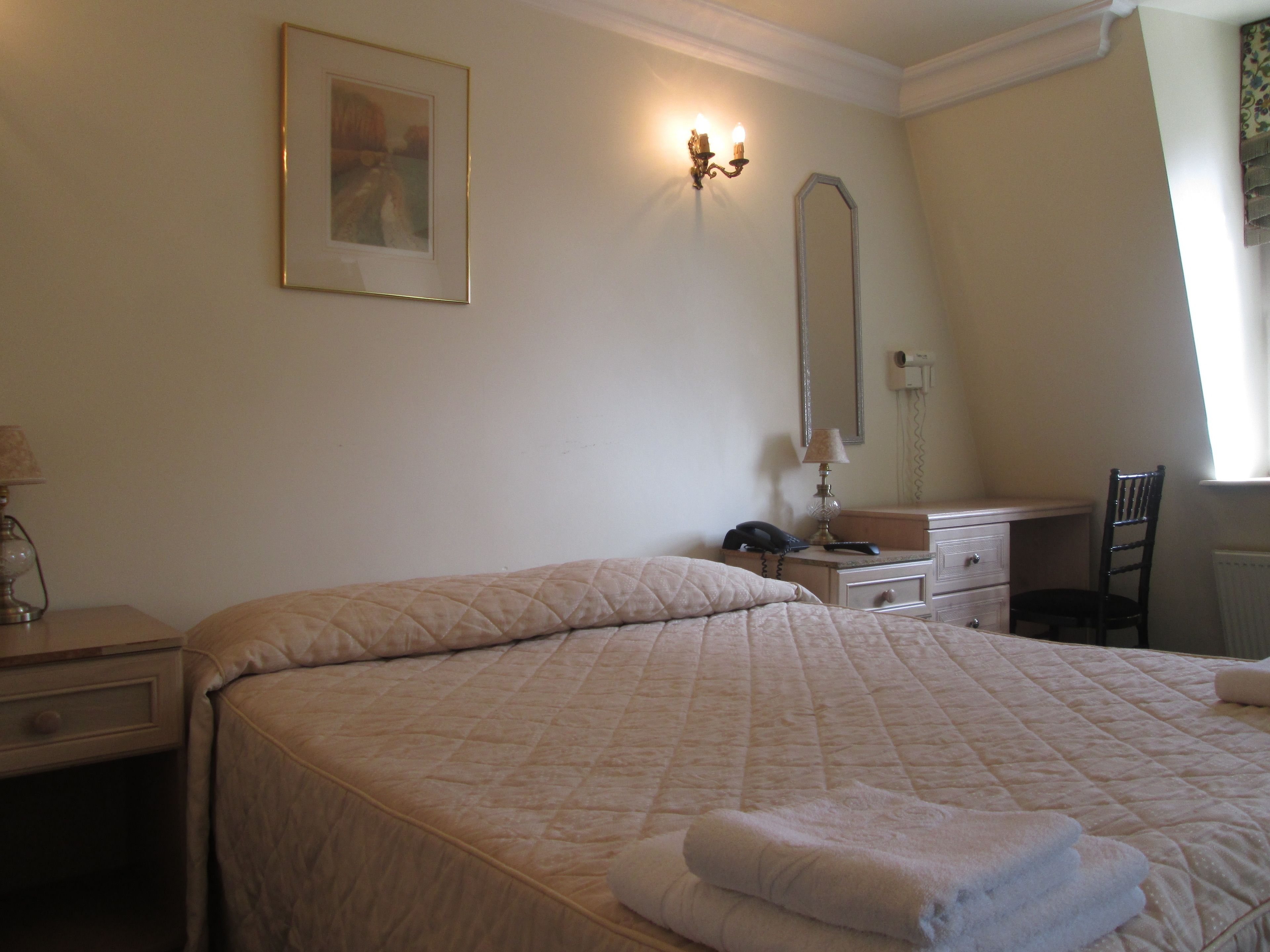 A hotel room with a large bed, a desk, a chair, a lamp, a telephone, and a mirror.