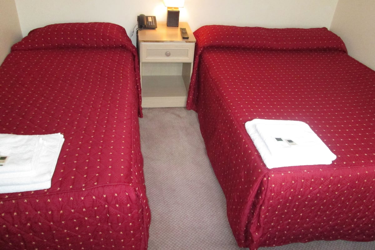 A hotel room with two single beds, each with a red bedspread and a white towel. There is a nightstand with a lamp and a phone between the beds.