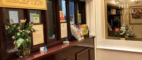 Un comptoir d’accueil à l’hôtel Brompton, avec un panneau indiquant la zone de réception, un panneau interdisant de fumer et des brochures d’information.