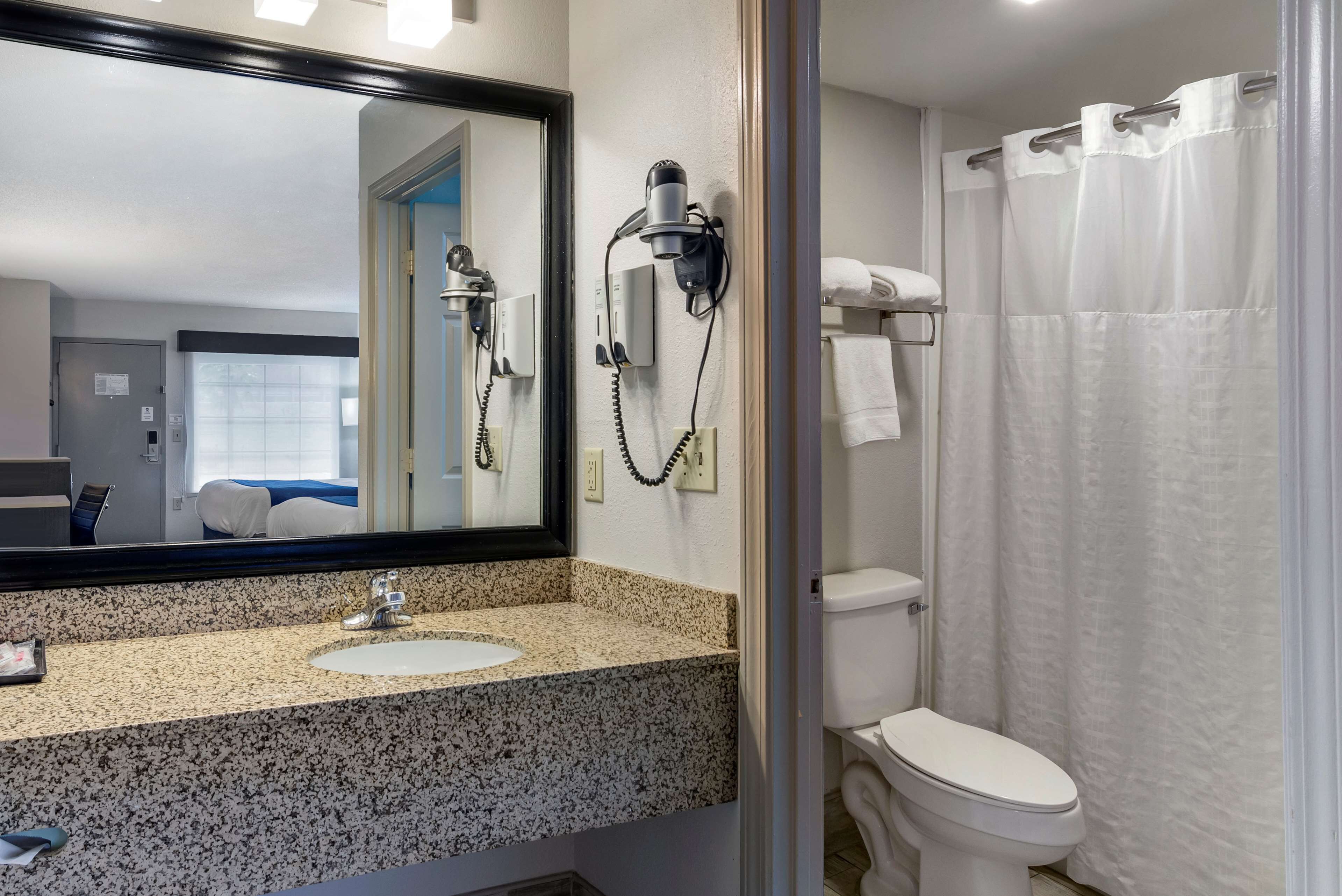 Un baño con lavamanos de encimera de granito, un secador de pelo sujeto a la pared, un inodoro y una cortina de ducha.