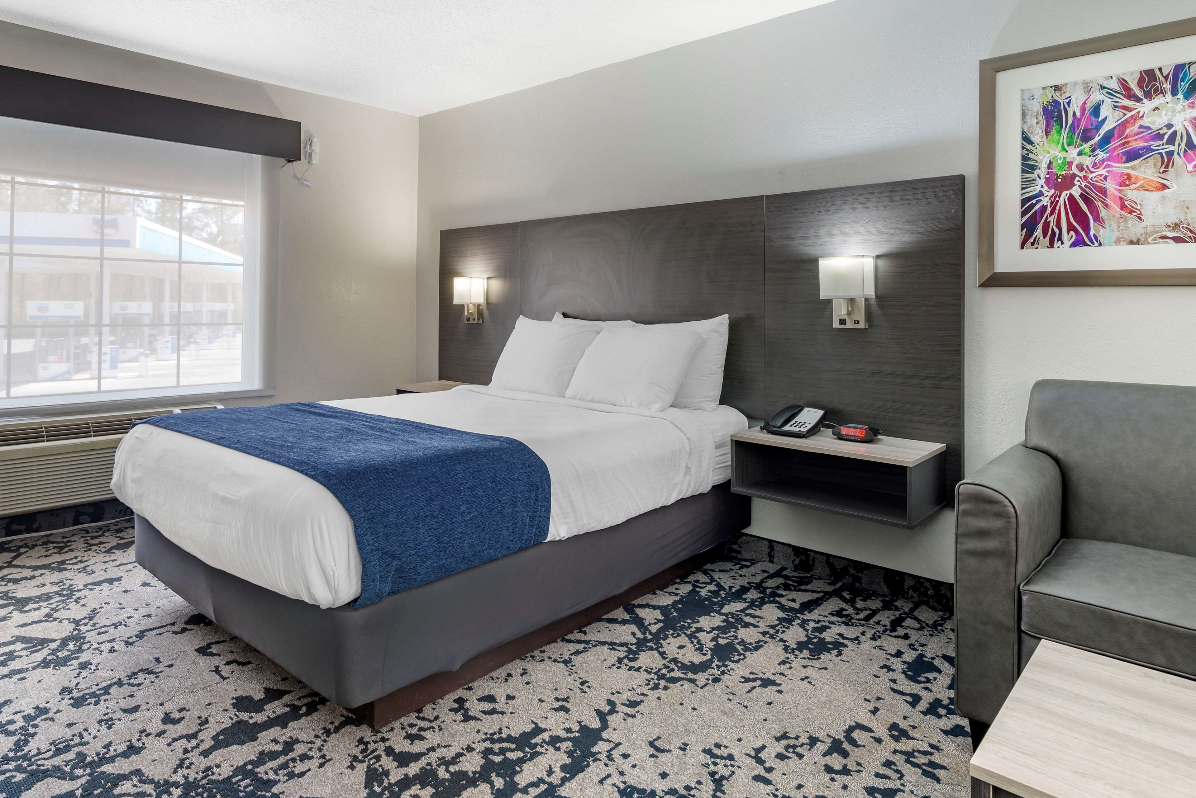A hotel room with a large bed, bedside lamps, a desk with a phone, a gray armchair, and a window with a view.