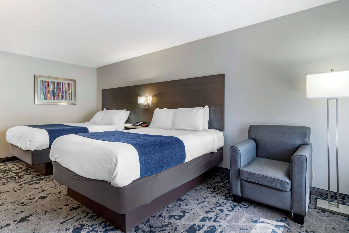 A hotel room with two beds, a grey armchair, a wall-mounted lamp, and a framed abstract painting.