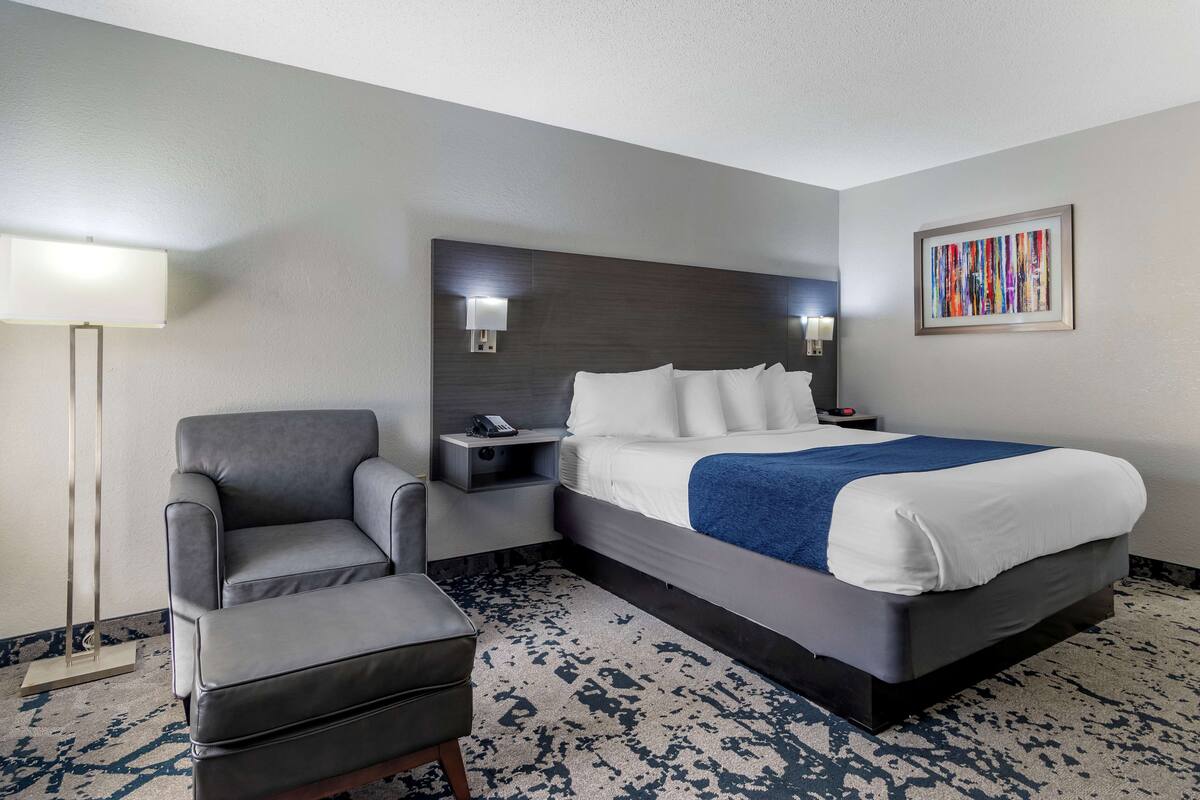 A hotel room with a large bed, a gray armchair, a bedside table with a lamp, and a wall-mounted artwork.