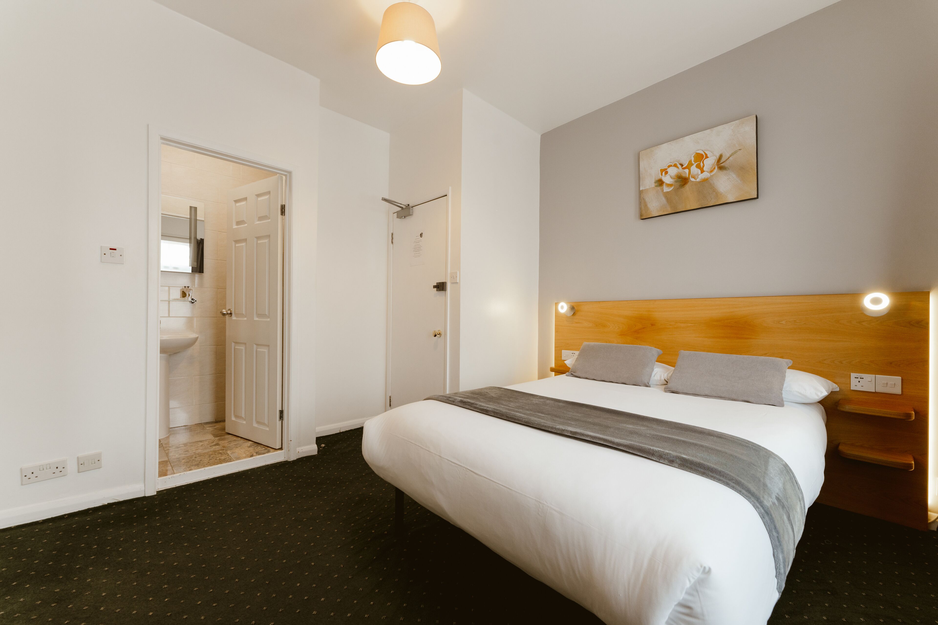 A hotel room with a bed, a wooden headboard, a wall-mounted light, and a door leading to a bathroom.
