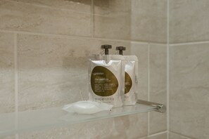 A bathroom with beige tiles, a glass shelf holding a white bottle and a white pouch, and a faucet. - Star Hotel (London)