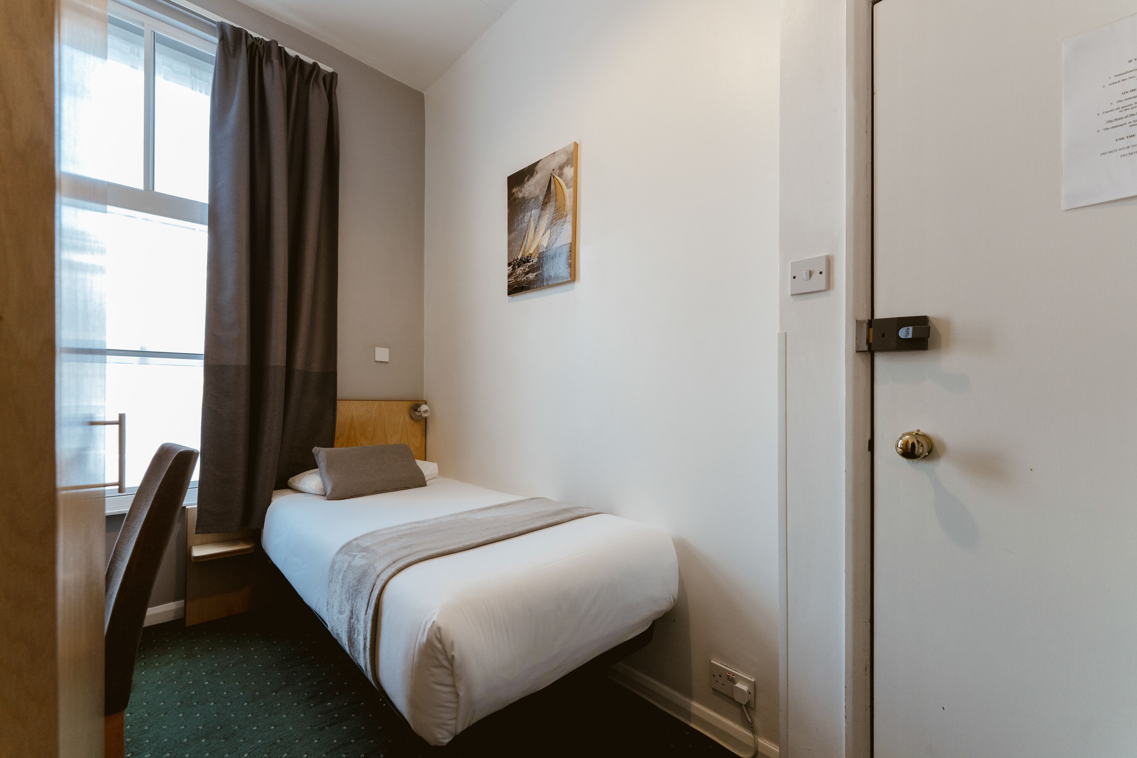 Een hotelkamer met een bed, een bureau, een stoel, een raam met gordijnen, een schilderij aan de muur en een deur met slot.
