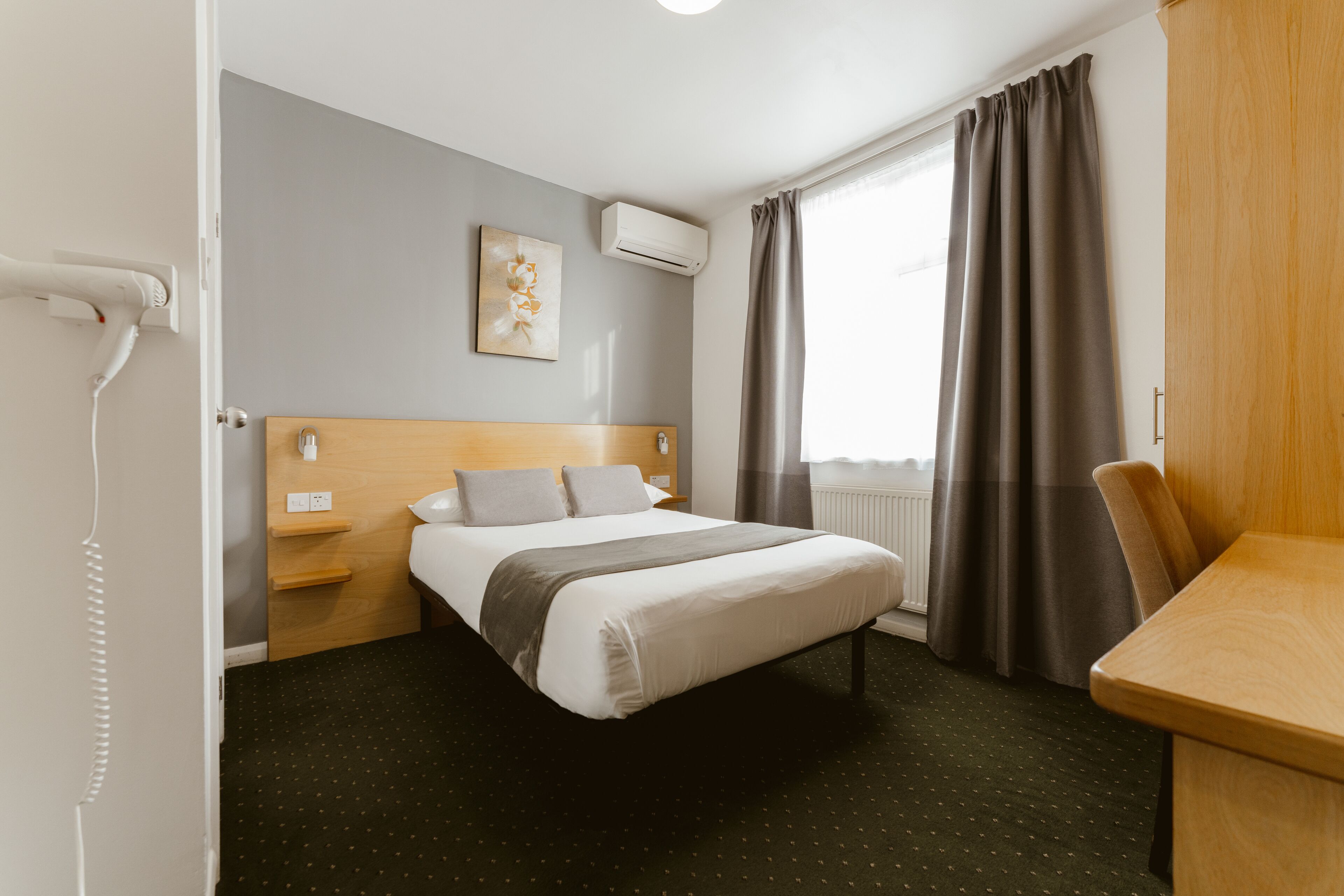 Una habitación de hotel con cama, escritorio, silla, aire acondicionado, ventana con cortinas y un cuadro en la pared.