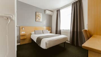 Una habitación de hotel con cama, escritorio, silla, aire acondicionado, ventana con cortinas y un cuadro en la pared.