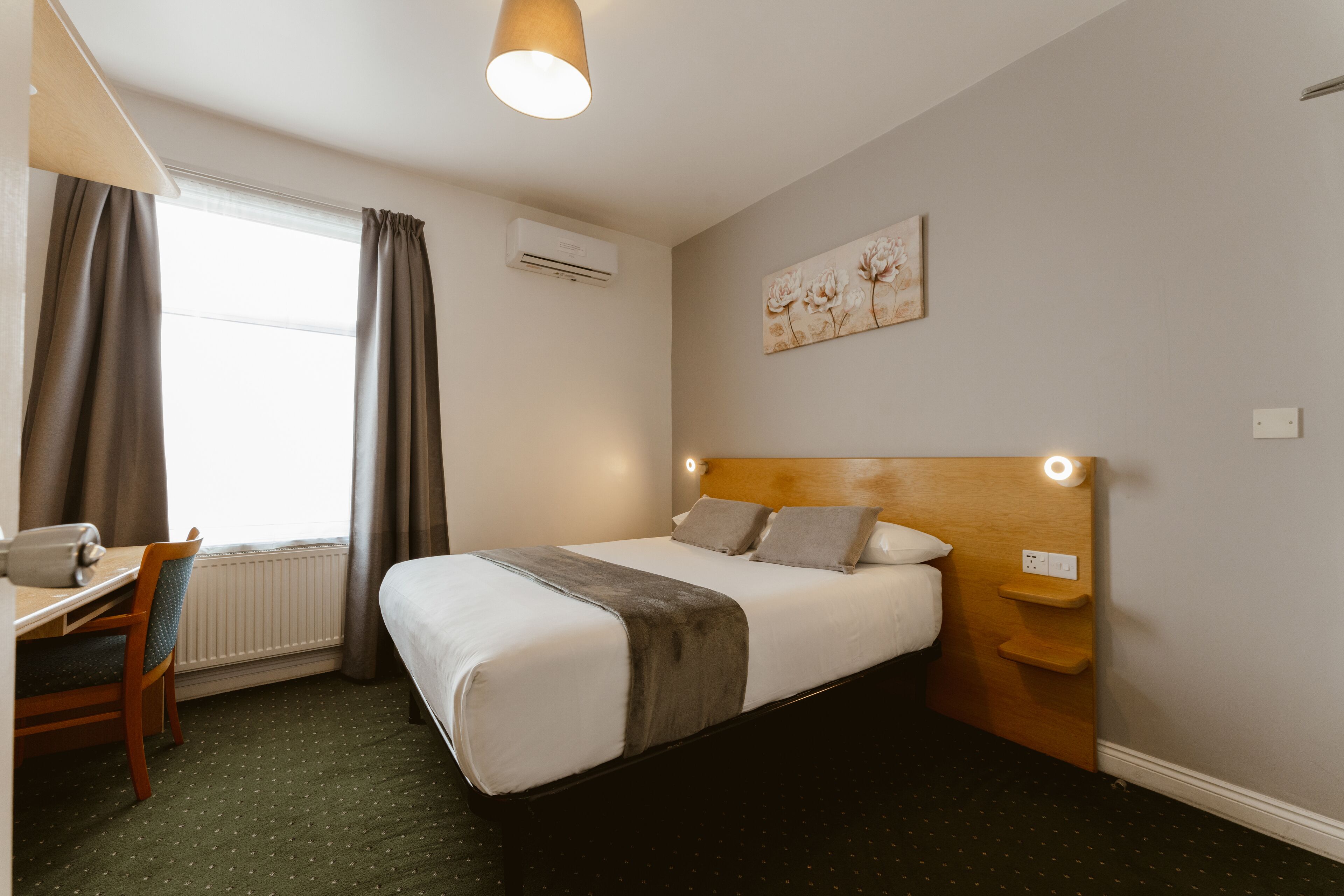 A hotel room with a bed, a desk, a chair, a window with curtains, an air conditioning unit, a wall-mounted artwork, and a wooden headboard with built-in lighting.